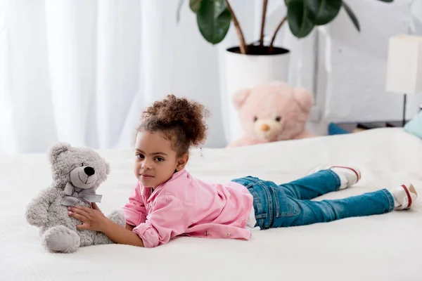 Curly african american kid lying on the bed with her teddy bear — Stock Photo