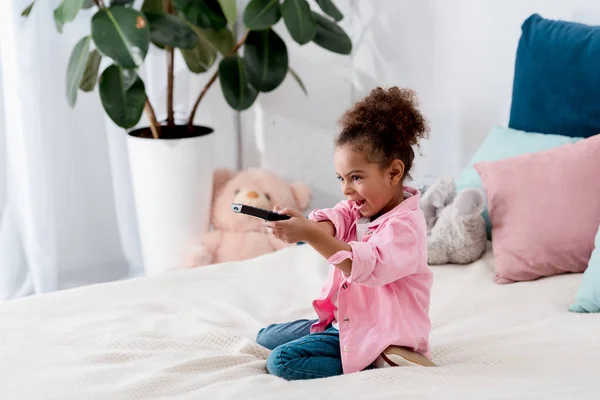 Criança afro-americana emocional sentada na cama com TV remota e trocando os canais — Fotografia de Stock