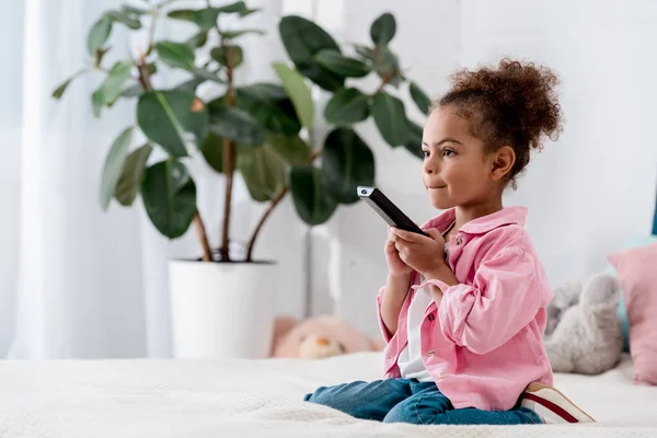 Criança afro-americana encaracolada sentada na cama e assistindo TV com controle remoto em suas mãos — Fotografia de Stock