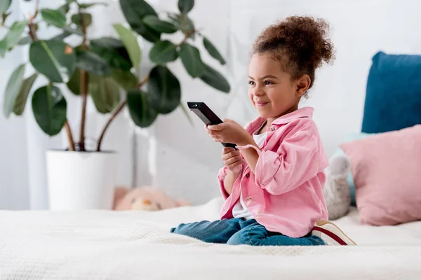 Emocionado niño afroamericano sentado en la cama con control remoto de televisión - foto de stock