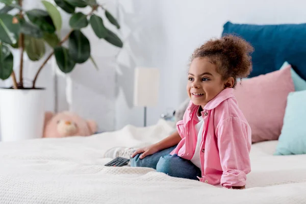 Sonriente niño afroamericano sentado en la cama y viendo el programa de televisión - foto de stock