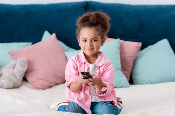 Enfant afro-américain bouclé assis sur le lit et appuyez sur les boutons de la télécommande — Photo de stock