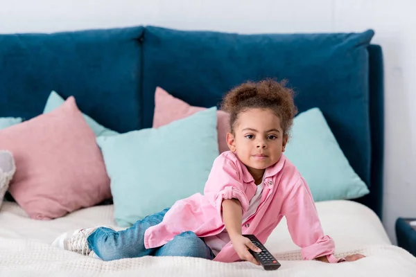 Süße afrikanische amerikanische Kind sitzt auf dem Bett mit TV-Fernbedienung — Stockfoto