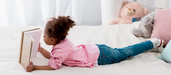 Entzückendes afrikanisch-amerikanisches Kind, das auf dem Bett liegt und ein Buch liest — Stockfoto