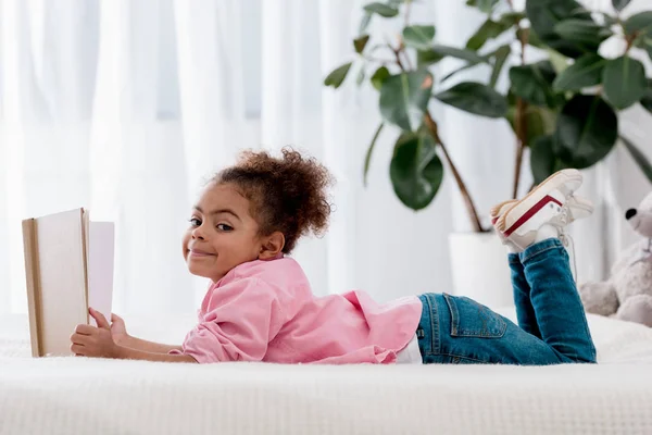 Vista lateral de un adorable niño afroamericano acostado en la cama y leyendo el libro - foto de stock