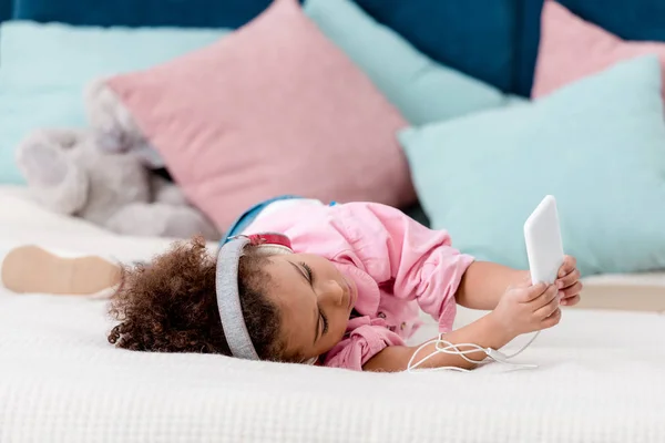 Adorable african american child lying on the bed and listening music on her smartphone in headphones — Stock Photo