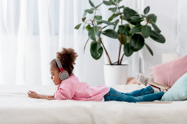 Vista lateral da linda criança afro-americana deitada na cama e ouvir música em seu smartphone em fones de ouvido — Fotografia de Stock