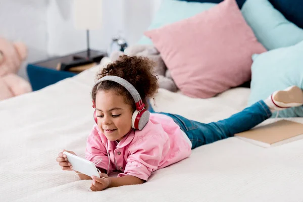 Mignon afro-américain enfant couché sur le lit et écouter de la musique sur son smartphone dans les écouteurs — Photo de stock
