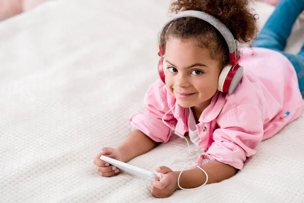 Niño afroamericano sonriente acostado en la cama y escuchando música en su teléfono inteligente con auriculares - foto de stock