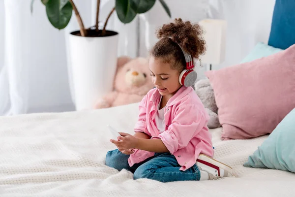 Niño afroamericano reflexivo sentado en la cama en auriculares y con teléfono inteligente - foto de stock
