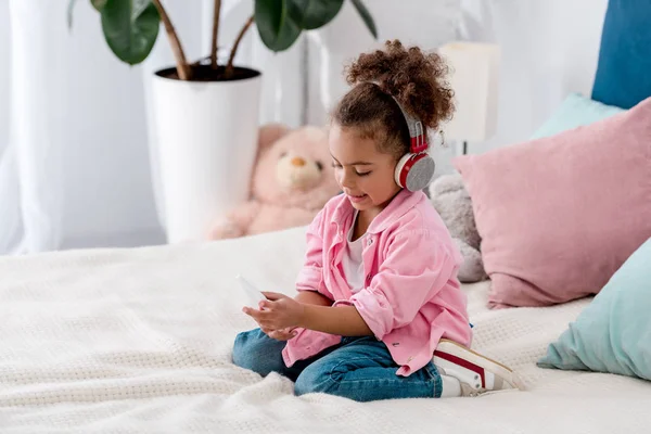 Thoughtful african american kid sitting on the bed in headphones and with smartphone — Stock Photo