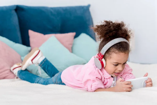 Adorable afroamericano niño acostado en la cama y escuchar música en los auriculares - foto de stock