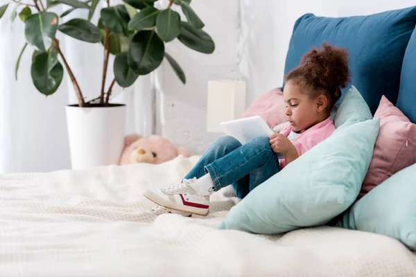 Lockiges afrikanisch-amerikanisches Kind sitzt mit digitalem Tablet auf dem Bett — Stockfoto