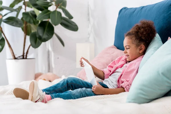 Lächelnd afrikanisches amerikanisches Kind auf dem Bett liegend mit digitalem Tablet — Stockfoto