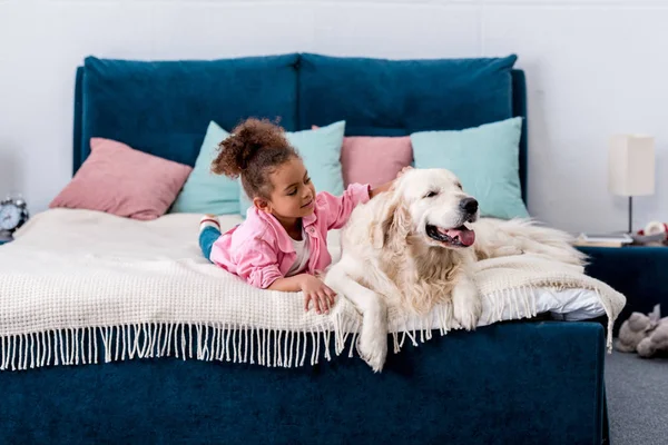 Adorabile bambino afroamericano sdraiato sul letto e che gratta la schiena di un cane — Foto stock