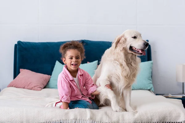 Sorrindo afro-americano criança sentada na cama e segurando a pata do cão — Fotografia de Stock