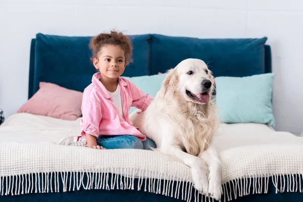 Cute african american child sitting on the bed with her dog beside — Stock Photo