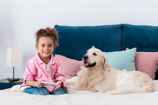 Smiling african american kid sitting on the bed with smartphone and golden retriever — Stock Photo