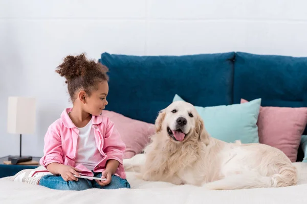 Criança americana africana encaracolada sentada na cama com smartphone e sorrindo golden retriever — Fotografia de Stock