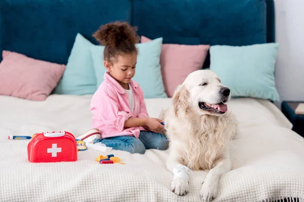 Bonito criança afro-americana sentada na cama com brinquedos perto de seu cão — Fotografia de Stock