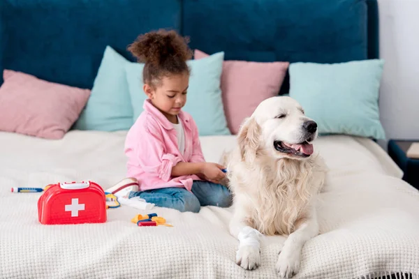 Süße afrikanisch-amerikanische Kind sitzt auf dem Bett mit Spielzeug in der Nähe ihres Hundes — Stockfoto