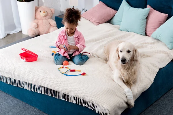 Bonito criança afro-americana brincando com brinquedos na cama com cão branco deitado ao lado — Fotografia de Stock