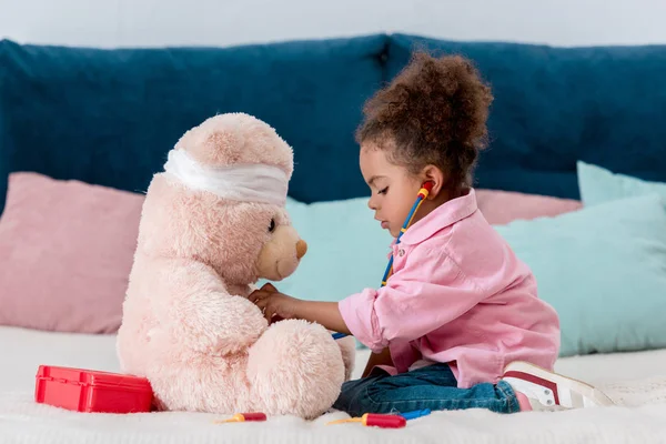 Little african american child in pink jacket playing the doctor  with teddy bear — Stock Photo