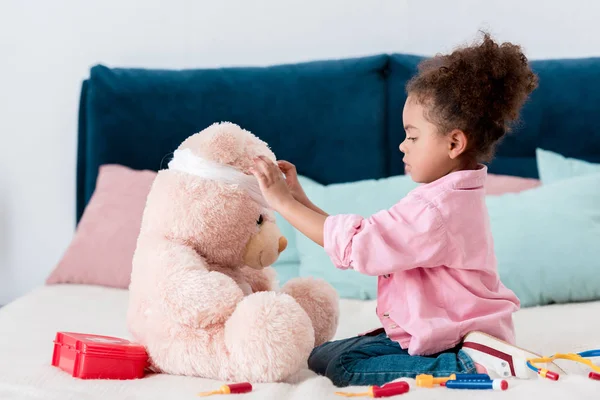 Piccolo bambino afroamericano in giacca rosa che gioca al dottore con l'orsacchiotto — Stock Photo