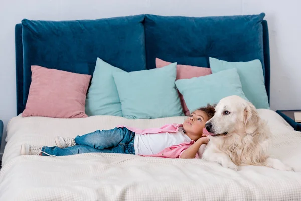 Criança americana africana adorável em jaqueta rosa deitada na cama com o cão enquanto olha para longe — Fotografia de Stock