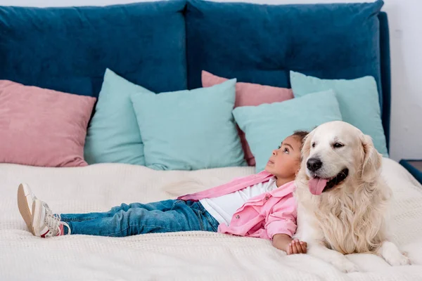 Adorable little african american kid in pink jacket lying on bed and leaning to dog — Stock Photo