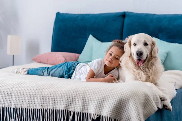 Bonito menino afro-americano pequeno com cão feliz deitado na cama com almofadas coloridas — Fotografia de Stock