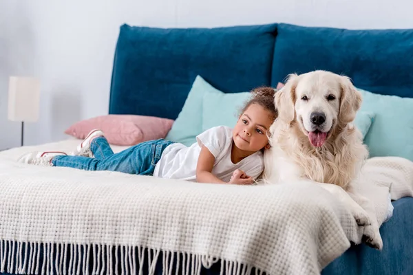 Adorable niño afroamericano con perro feliz mirando hacia otro lado mientras está acostado en la cama - foto de stock