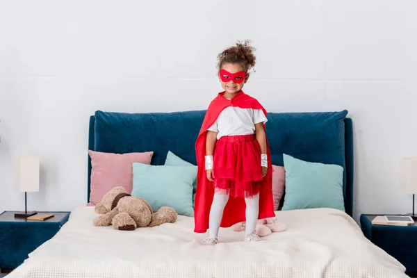 Petit enfant afro-américain souriant en costume de super-héros debout sur le lit avec des oreillers colorés — Photo de stock