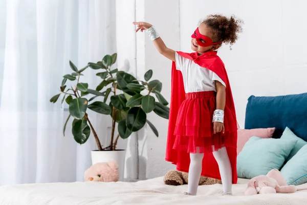 Entzückendes kleines afrikanisch-amerikanisches Kind im roten Superheldenkostüm gestikuliert, während es auf dem Bett steht — Stockfoto