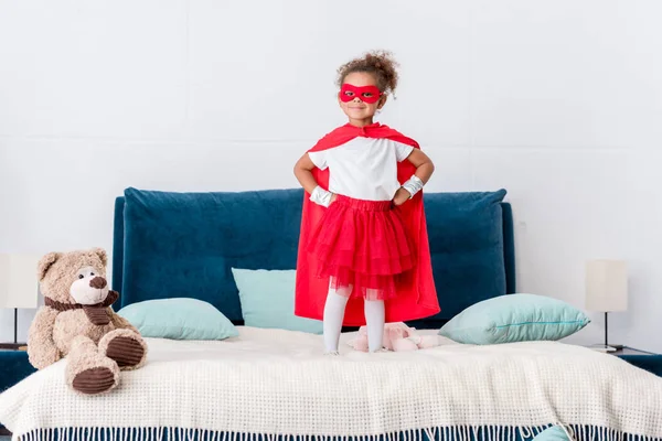 Little african american kid in red superhero costume and mask with hands on hips standing on bed — Stock Photo