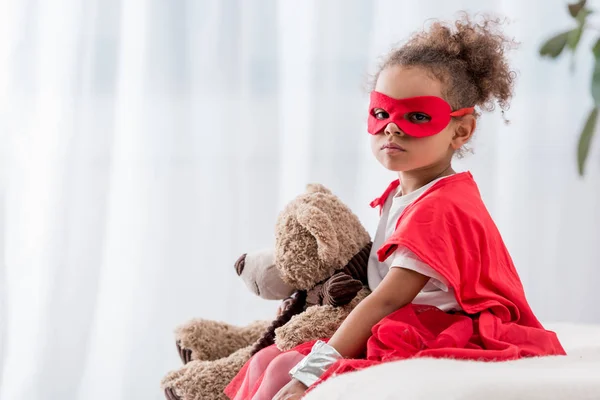 Cute african american child in red superhero costume and mask with teddy bear looking at camera — Stock Photo