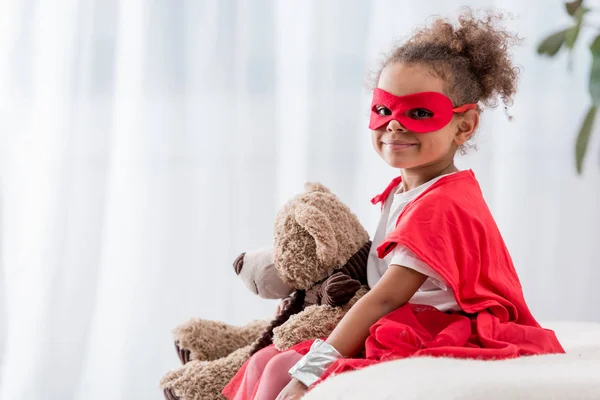Adorable petit enfant afro-américain en costume de super-héros et masque avec ours en peluche souriant à la caméra — Photo de stock