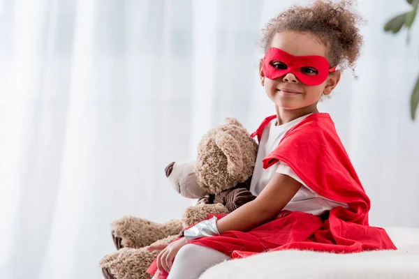 Adorable niño afroamericano en traje de superhéroe y máscara con oso de peluche - foto de stock