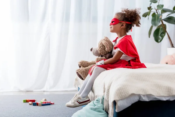 Vista lateral del niño afroamericano en traje de superhéroe y máscara con oso de peluche - foto de stock