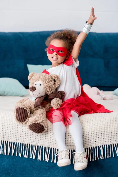 Adorável pequena criança afro-americana em traje de super-herói sentado na cama com ursinho de pelúcia — Fotografia de Stock