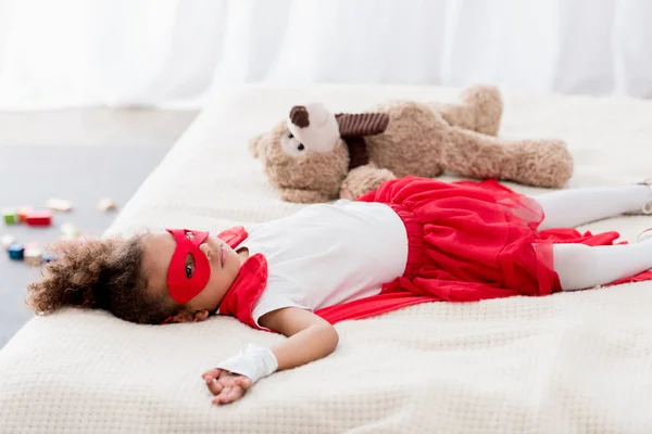 Bonito pequena criança afro-americana em traje de super-herói e máscara deitada na cama com ursinho de pelúcia — Fotografia de Stock