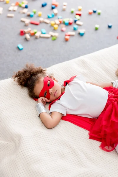 Cute little african american child in red superhero costume and red mask lying on bed — Stock Photo