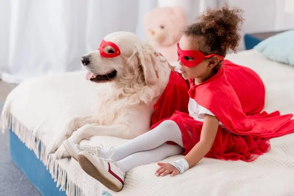 Adorable little african american kid with dog in red superhero costumes sitting on bed — Stock Photo