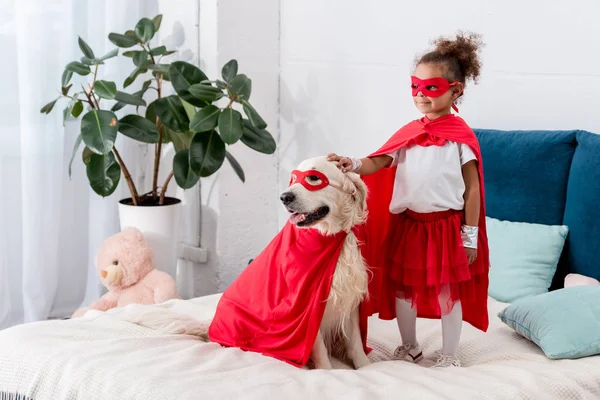 Cute little african american kid with dog in superhero costumes looking away while standing on the bed — Stock Photo