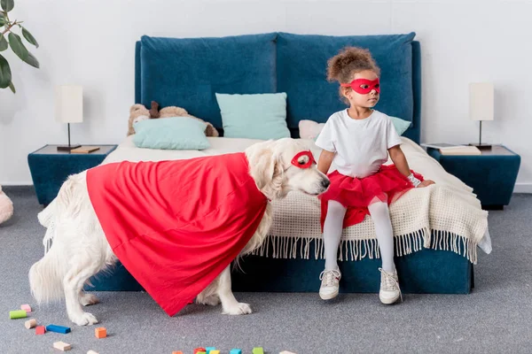 Cute little african american kid with dog in superhero costumes in bedroom — Stock Photo