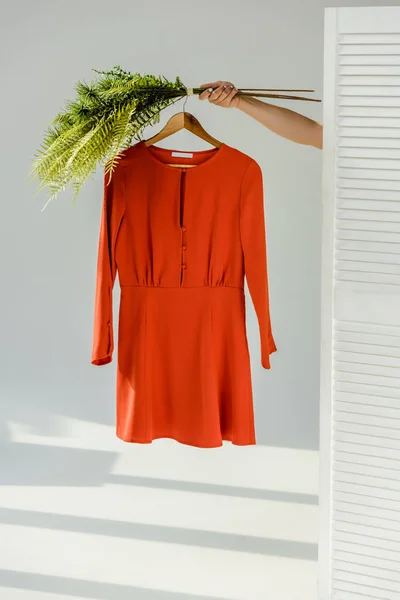 Female hand holding hanger with red stylish dress and green plants near room divider — Stock Photo