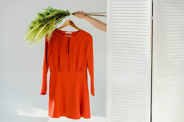 Mão feminina segurando cabide com vestido elegante vermelho e plantas verdes perto divisor de quarto — Fotografia de Stock