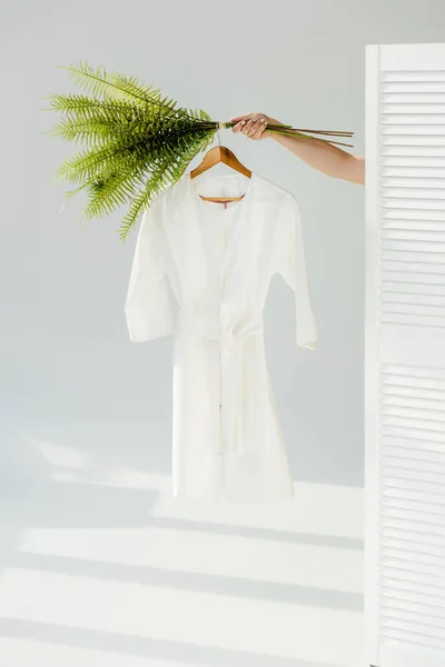 Mão feminina segurando cabide com vestido elegante branco e plantas verdes — Fotografia de Stock