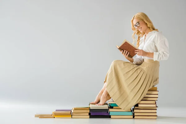 Attractive smiling barefoot woman in glasses sitting on steps made of books — Stock Photo