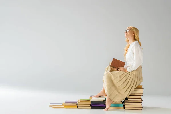 Attractive dreaming barefoot woman in glasses sitting on steps made of books — Stock Photo